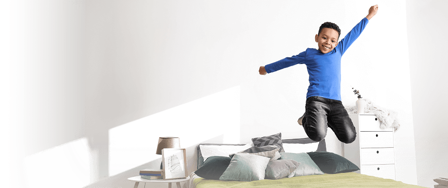 Young boy in a blue shirt joyfully jumping on a bad in a bright, modern bedroom